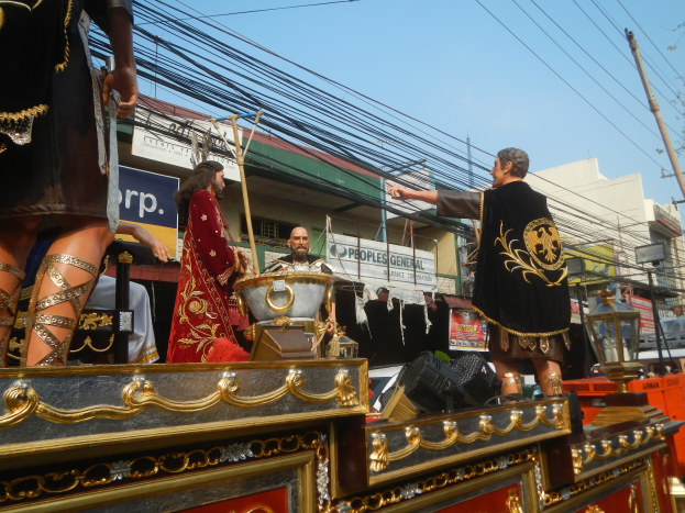 Eine Gruppe von Menschen in Kostümen fährt auf einem hell erleuchteten Paradewagen, mit Gebäuden, Schildern, Pfählen, Drähten, Bäumen und einem klaren blauen Himmel im Hintergrund.