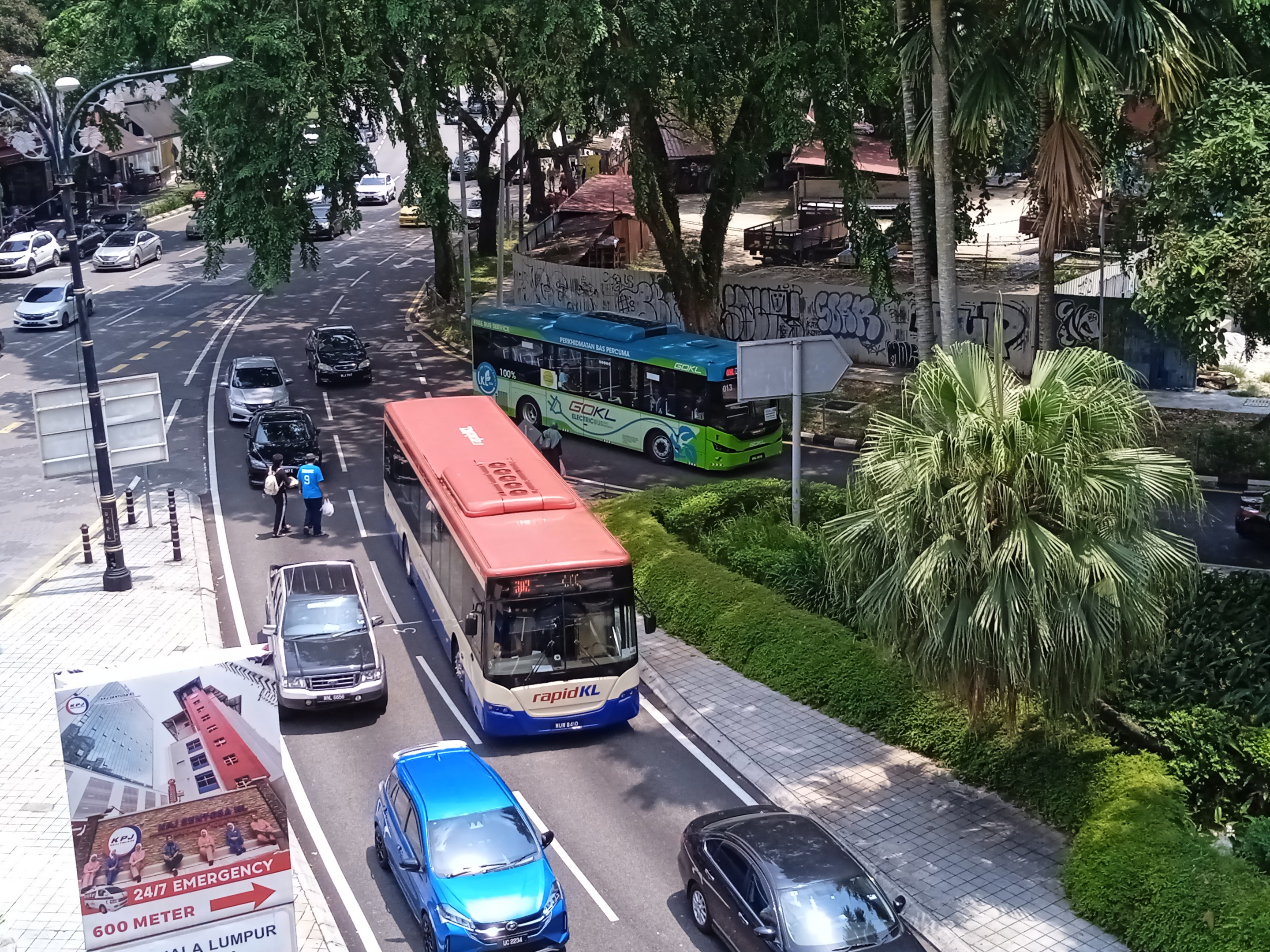 Eine belebte Stadtstraße mit mehreren Autos und Bussen, beleuchtet von Straßenlaternen, Fußgänger auf dem Gehweg, Grünzeug auf der rechten Seite und Gebäude im Hintergrund.