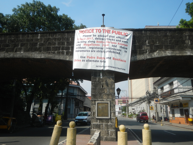 Eine Brücke mit einem "Hinweis an die Öffentlichkeit"-Schild, Fahrzeuge auf der Straße darunter und Gebäude, Bäume, Pfähle, Drähte und einen klaren blauen Himmel im Hintergrund.