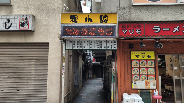 Eine enge Gasse, flankiert von Gebäuden mit Fensterläden, mit Schildern mit Text und Bildern, die zu einer Straße am Ende führt.
