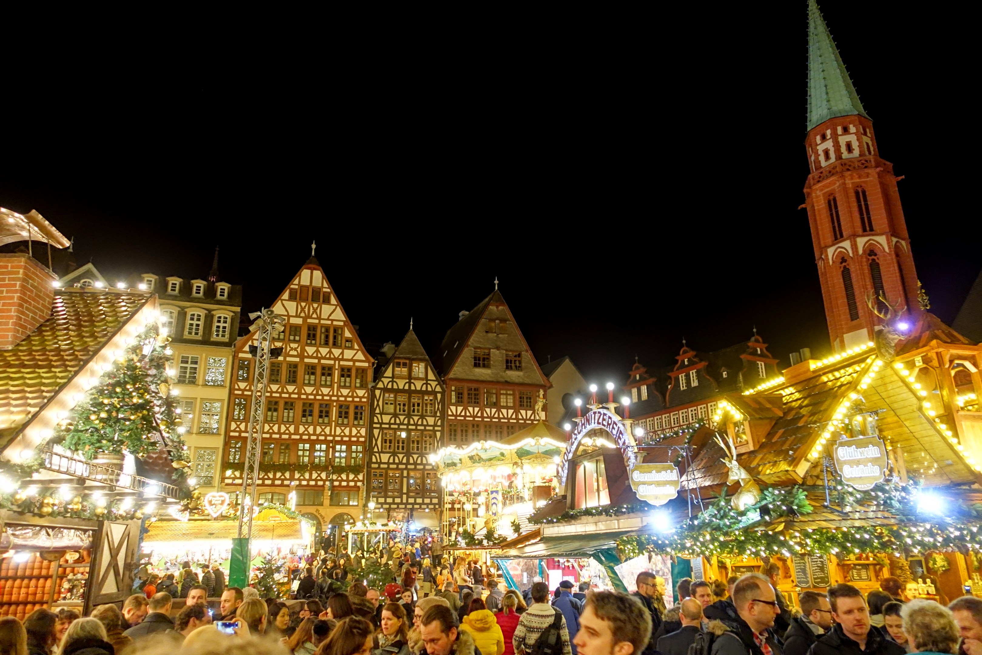 Ein geschäftiger Weihnachtsmarkt in Nürnberg, Deutschland mit Menschen um geschmützte Stände, festliche Lichter, Gebäude mit Fenstern, einen Uhrenturm und einen dunklen Himmel.
