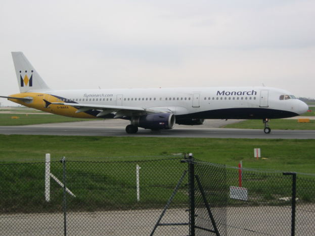 Ein Monarch Airlines Airbus A320-200 auf einer von Gras umgebenen Rollbahn, einem Zaun mit Pfählen und Brettern, Bäumen und einem klaren blauen Himmel.