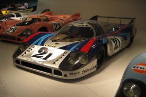 Ein Porsche 917K Le Mans Rennwagen in einem Museum ausgestellt, umgeben von anderen Sportwagen, mit Menschen in der Nähe und einer Wand im Hintergrund.