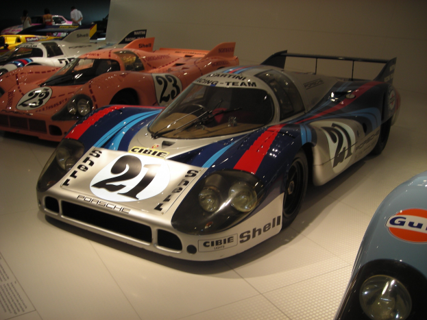 Ein Porsche 917K Le Mans Rennwagen in einem Museum ausgestellt, umgeben von anderen Sportwagen, mit Menschen in der Nähe und einer Wand im Hintergrund.
