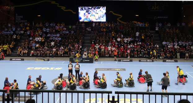 Eine Gruppe von Rollstuhlfahrern bei den Invictus Games Düsseldorf 2020, mit einer großen Zuschauermenge hinter einem Geländer, Treppen, die zu einem Bildschirm führen, und Informationsschildern um den Platz.