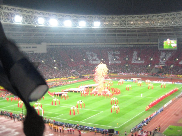 Ein Stadion mit Flutlicht, Eisengittern, Zuschauern, stehenden Personen, Werbetafeln, Bildschirmen, Überwachungskamera, Fahnen und Dekorationen.