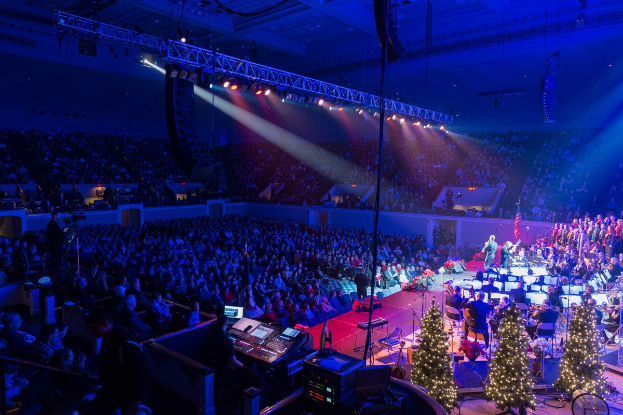 Eine große Menschenmenge in einem geschmückten Auditorium schaut einer Band zu, die auf einer Bühne mit Weihnachtsbäumen, Lautsprechern und festlicher Beleuchtung spielt.