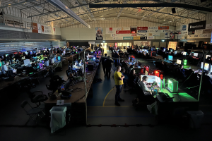 Ein großer Raum mit Menschen, die Videospiele spielen, sitzend und stehend, umgeben von Monitoren, Tastaturen, Mäusen und Kabeln auf Tischen, mit markierten Platten im Hintergrund und einer sichtbaren Decke.