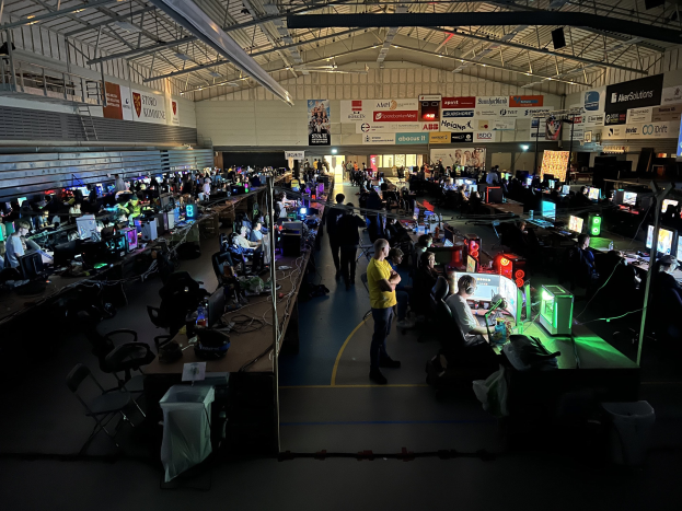 Ein großer Raum mit Menschen, die Videospiele spielen, sitzend und stehend, umgeben von Monitoren, Tastaturen, Mäusen und Kabeln auf Tischen, mit markierten Platten im Hintergrund und einer sichtbaren Decke.