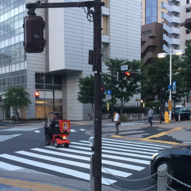Ein Roller ist auf der Straße mit einigen Personen im Hintergrund, eine Ampel im Vordergrund und Gebäude mit Bäumen in der Ferne.
