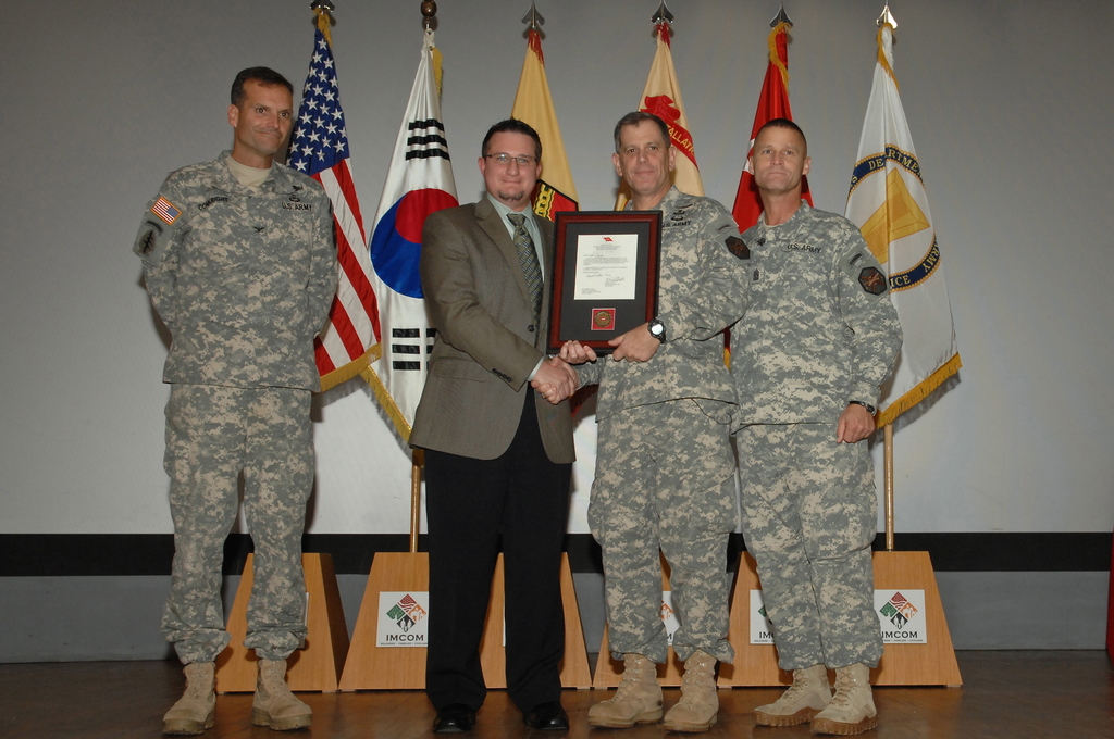 Vier Männer stehen auf einer Bühne: drei in Militäruniformen und einer in Blazer, Krawatte und Hemd, die Hände schütteln und einen Preis überreichen, mit sechs Fahnen an Stangen und einer weißen Wand im Hintergrund.
