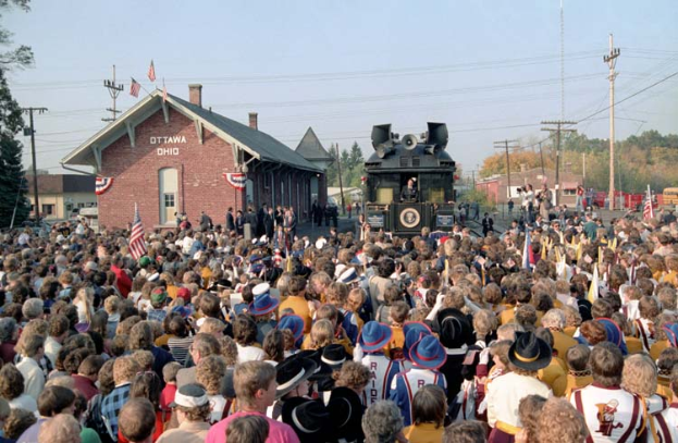 Eine Menschenmenge steht vor einem Zug auf Schienen, einige tragen Hüte und halten Fahnen, während Strommasten und -leitungen neben dem Zug verlaufen, mit Gebäuden, Bäumen und einem klaren blauen Himmel im Hintergrund.