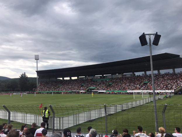 Eine große Menschenmenge schaut einem Fußballspiel in einem Stadion zu, das von einem Zaun umgeben ist, mit Laternenmasten, die das Feld beleuchten, Bäumen und Hügeln im Hintergrund und einer bewölkten Himmel.