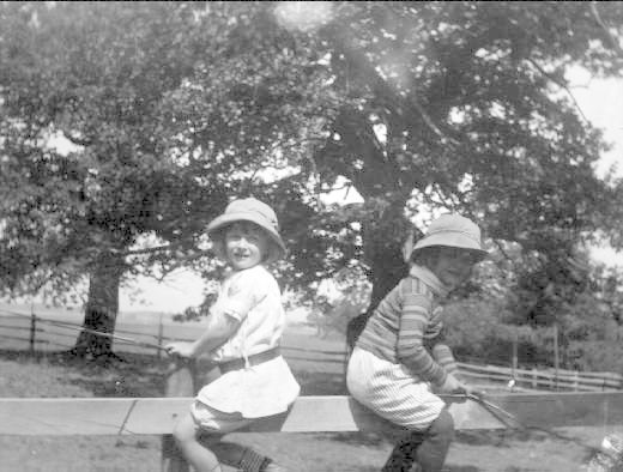 Zwei junge Jungs in Mützen und Baseball-Uniformen stehen in einem grünen Park und halten Schläger, mit Bäumen und einem Zaun im Hintergrund, auf einem Schwarz-Weiß-Foto.