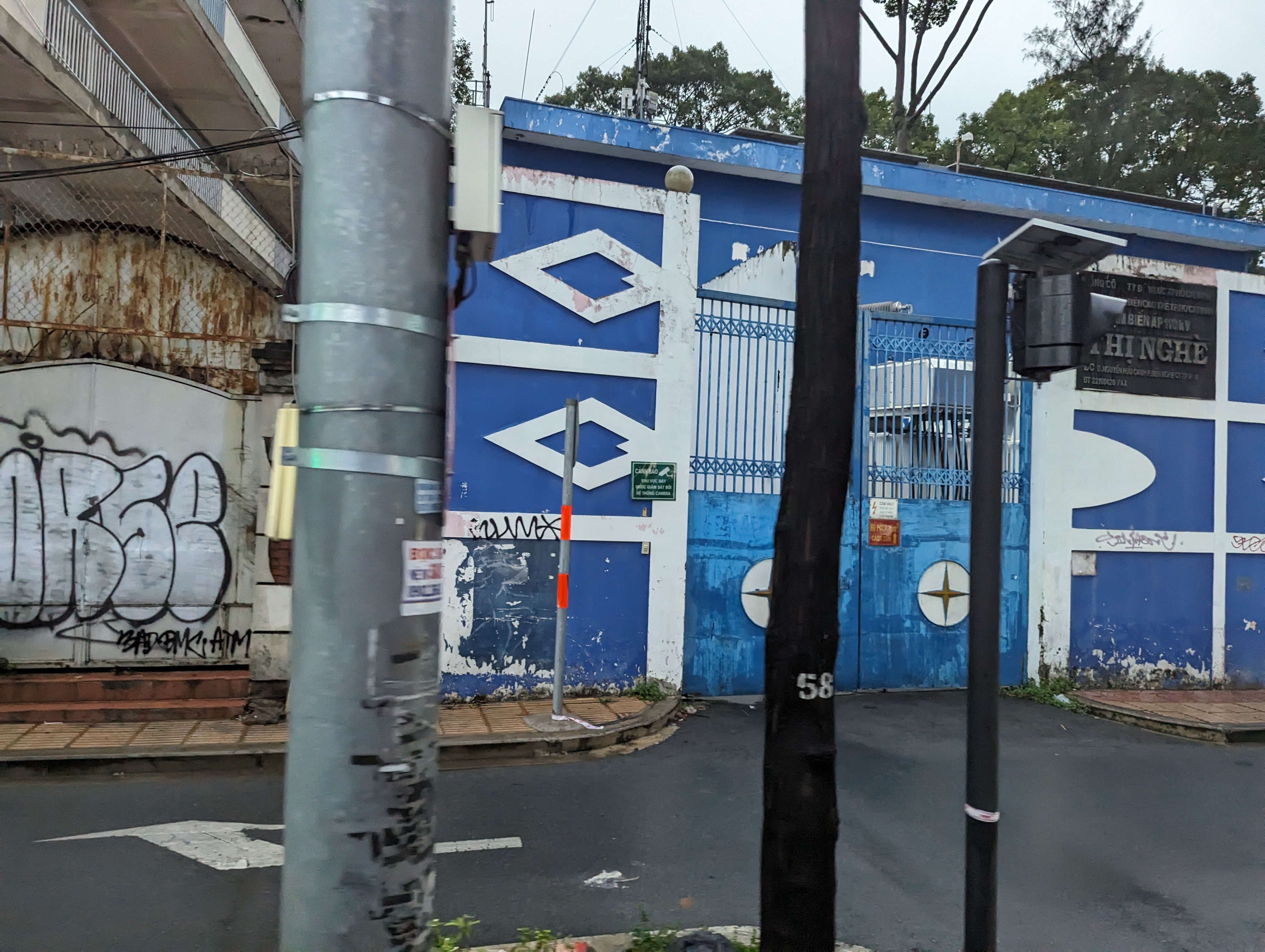 Ein blaues Gebäude mit Graffiti, ein Metallzaun, eine Baumgruppe, mehrere Pfosten, eine Ampel, Drähte und ein bewölkter Himmel.
