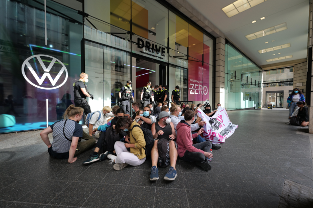 Eine Gruppe von Menschen sitzt vor einer Volkswagen-Vertretung mit Glaswänden und einem Logo auf der linken Seite auf dem Boden, einige halten ein Transparent.