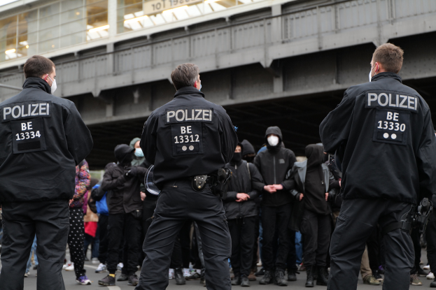 Eine Gruppe von Polizisten in schwarzen Uniformen und Masken steht vor einer Menge, mit einer Brücke und einem Gebäude im Hintergrund, während einer Demonstration in einer Stadt.