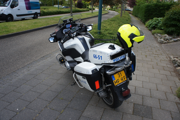 Eine Polizeimotorrad auf dem Seitenstreifen mit einem Helm darauf, umgeben von Bäumen, Pfählen, Gebäuden, Pflanzen, Gras, Steinen und anderen Fahrzeugen.