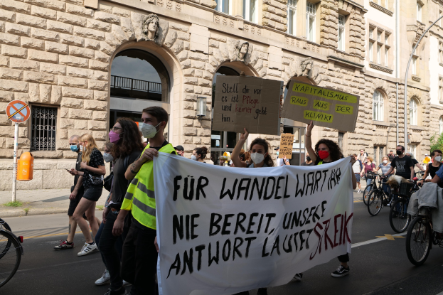 Eine Gruppe von Menschen, die auf einer Demonstration durch die Straße marschieren, einige halten Schilder und andere fahren Fahrräder, mit einem Gebäude mit Fenstern, Bögen, Säulen und Skulpturen im Hintergrund.