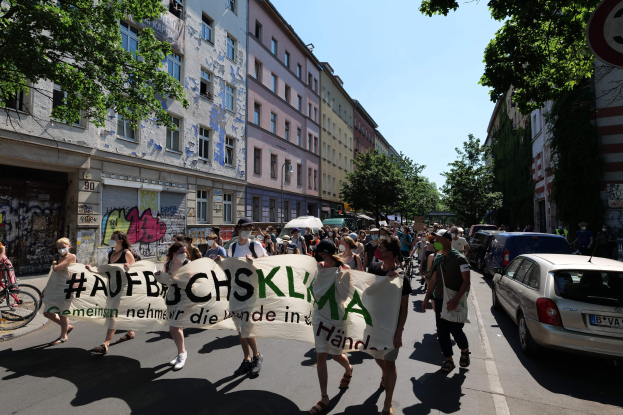 Eine Gruppe von Menschen bei einer Demonstration mit einem Transparent, Autos auf der rechten Seite und einem Radfahrer auf der linken Seite, vor dem Hintergrund von Gebäuden, Bäumen und einem Schild in Berlin.