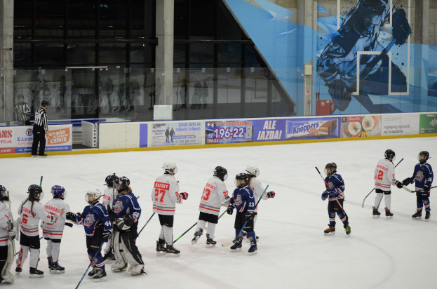 Gruppe junger Menschen, die Eisockey auf einer Indoor-Eisfläche spielen, mit Helmen und Stöcken, während eine Person links steht und Banner, Säulen und Glaswände im Hintergrund zu sehen sind.