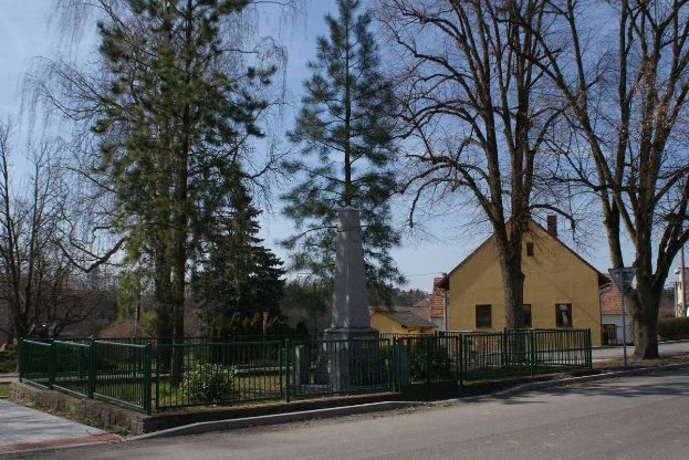 Denkmal für Holocaust-Opfer in der Mitte einer Straße, umgeben von einem Metallzaun mit Grünfläche, Häusern, einem Straßenmast und einem bewölkten Himmel.