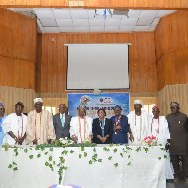 Gruppe von Menschen um einen weißgedeckten Tisch versammelt, der mit Blumen und Blättern geschmückt ist, bei der ICCI International Trade Fair 2019, mit einem Banner, Projektionsbildschirm und Deckenbeleuchtung im Hintergrund.
