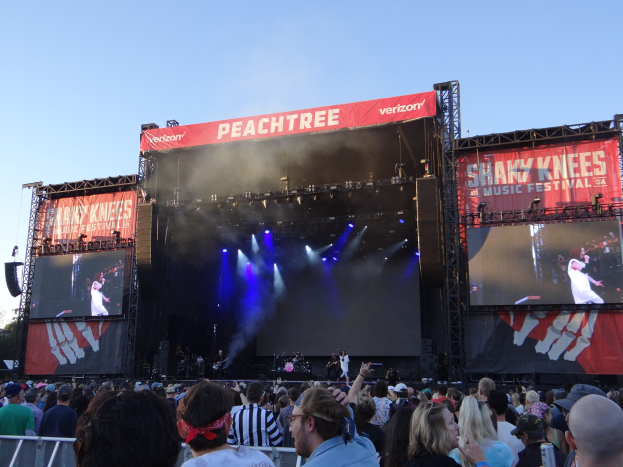 Eine große Menschenmenge steht vor einer hell beleuchteten Bühne auf dem Shaky Knees Music Festival, wo Musiker unter Bannern spielen, mit einem Geländer im Vordergrund und einem klaren blauen Himmel im Hintergrund.