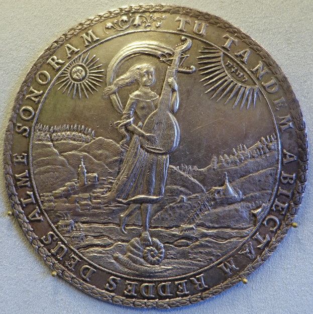 Eine Silbermedaille mit einer Frau in einem langen Kleid, die eine Harfe spielt, stehend in einem hohen Grasfeld unter einem klaren blauen Himmel, mit der Inschrift 'Medaille' darauf.