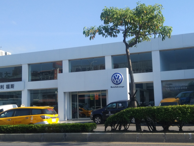 Ein Volkswagen-Händler mit Glaswänden und -fenstern, der Autos zeigt, die vor ihm geparkt sind, umgeben von Pflanzen und einem Baum unter einem sichtbaren Himmel.