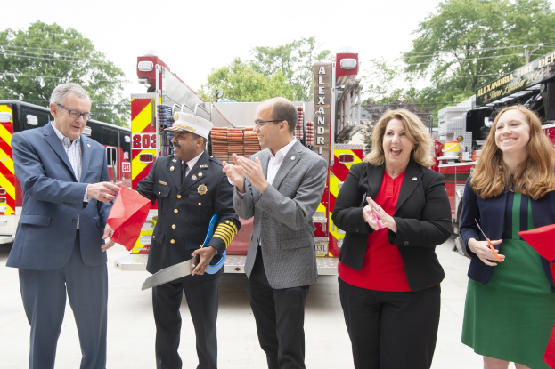 Eine Gruppe von Menschen steht vor einem Feuerwehrauto, klatscht und lacht, während zwei eine Schere und ein rotes Tuch halten, bei einer Eröffnungsfeier der Alexandria Fire Department, mit Bäumen, Drähten und einem klaren blauen Himmel im Hintergrund.