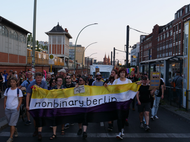 Gruppe von Menschen, die eine 'Nonbinary Berlin'-Fahne schwingend durch eine Berliner Straße gehen, mit Laternenpfählen, Gebäuden, einer Bushaltestelle, Bäumen und einem klaren blauen Himmel im Hintergrund.