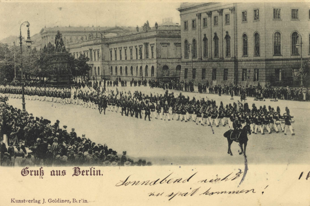 Ein altes Schwarz-Weiß-Foto eines Umzugs in Berlin, Deutschland, mit Menschen auf dem Boden und auf Pferden, einer Statue auf einem Podest, Straßenlaternen, Bäumen, Gebäuden mit Fenstern und einem bewölkten Himmel, mit Text am unteren Rand des Bildes.