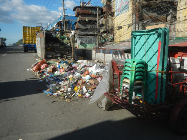 Ein Lastwagen neben einem Müllhaufen auf einer Straße geparkt, mit einem mit Plastikstühlen gefüllten Handwagen rechts daneben und Gebäuden, Strommasten, Bäumen und einem bewölkten Himmel im Hintergrund.