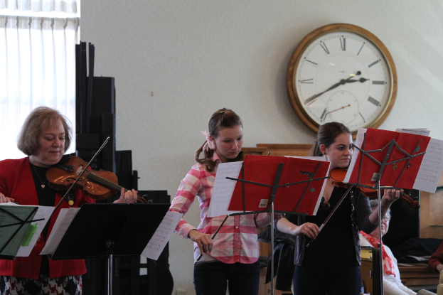 Drei Frauen auf der Bühne bei einem musikalischen Konzert, die Geigen spielen, mit Notenständern und Noten davor, während ein Zuschauer im Vordergrund sitzt; eine Uhr hängt an der weißen Wand im Hintergrund, und ein weißer Vorhang ist links zu sehen.