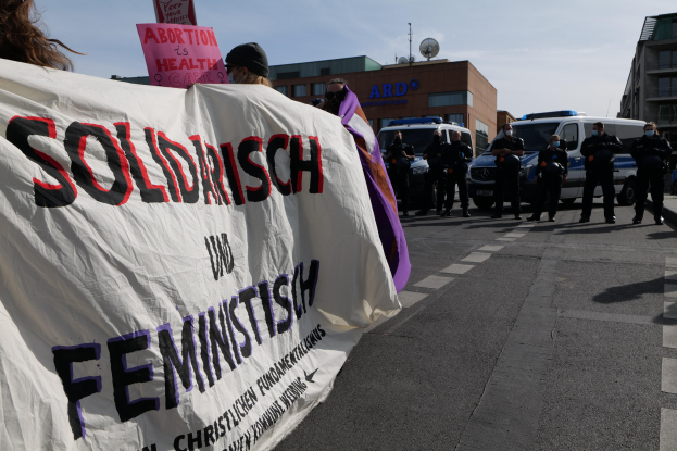Eine Gruppe von Menschen, die eine "Solidarität und Feminismus"-Fahne schwingt, marschiert die Straße entlang, mit geparkten Fahrzeugen, Gebäuden, einer Schüsselantenne und einem klaren blauen Himmel im Hintergrund.