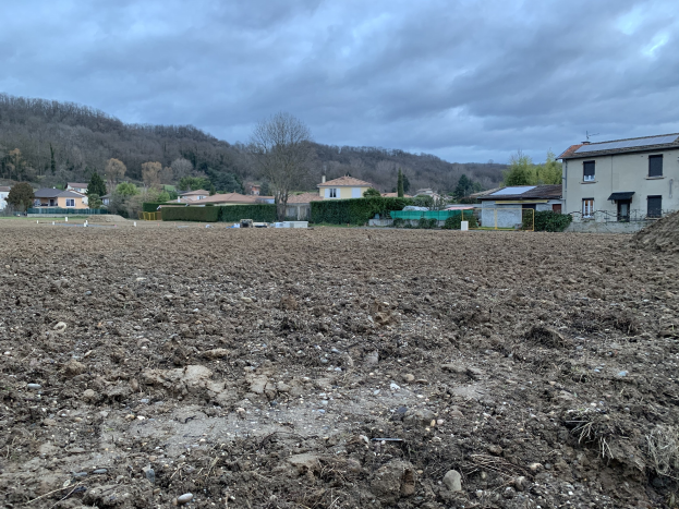 Großes freies Feld mit Erde, umgeben von Gebäuden, Vegetation und einem Zaun, mit Hügeln und bewölktem Himmel im Hintergrund, zum Verkauf angeboten zu 1.000.000€.