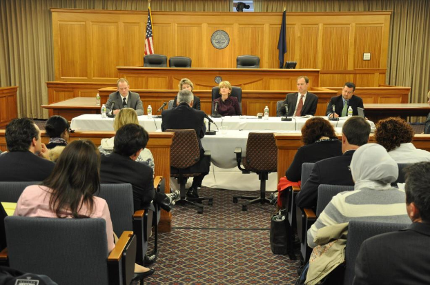 Tagungsraum mit einer Gruppe von Menschen, die auf Stühlen sitzen, zwei ausländischen Flaggen, einem orangen Schreibtisch mit Wasserflaschen und einem Mikrofon.