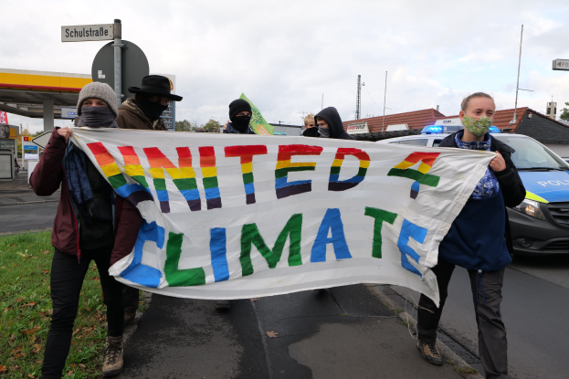 Eine Gruppe maskierter Personen hält ein 'United Climate'-Schild vor einer Tankstelle. Im Hintergrund sind Fahrzeuge, Gras, Schilder, Gebäude, Bäume und eine bewölkte Himmel zu sehen.