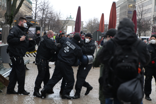 Eine Gruppe von Polizisten in schwarzer Einsatzkleidung und Masken, die eine Straße in Berlin, Deutschland, entlanggehen, mit Gebäuden, Bäumen, Schirmen, Pfählen, Fahrzeugen und einem sichtbaren Himmel im Hintergrund.