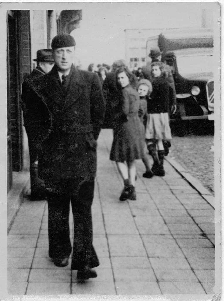 Ein Schwarz-Weiß-Bild, das mehrere Personen und ein Fahrzeug zeigt, wobei einige Individuals Hüte tragen und ein Gebäude in der Ferne zu sehen ist.