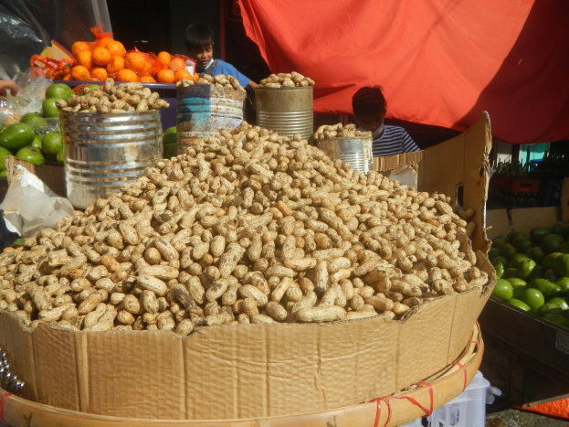 Korb mit Erdnüssen auf einem Tisch mit verschiedenen Früchten und Gemüse, zwei Kinder im Hintergrund, roter Stoff über den Tisch gelegt.