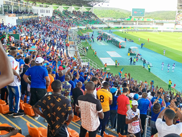 Eine große Menschenmenge versammelt sich vor einem eingezäunten Fußballfeld mit Hügeln und einem klaren blauen Himmel im Hintergrund und erwartet das Spiel der 2019 African Cup of Nations.