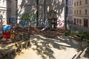 Stadtspielplatz mit Graffiti, umgeben von Gebäuden, Bäumen, Bänken, Pfosten, einem Mülleimer, Menschen und Fahrzeugen sowie sichtbarem Himmel.