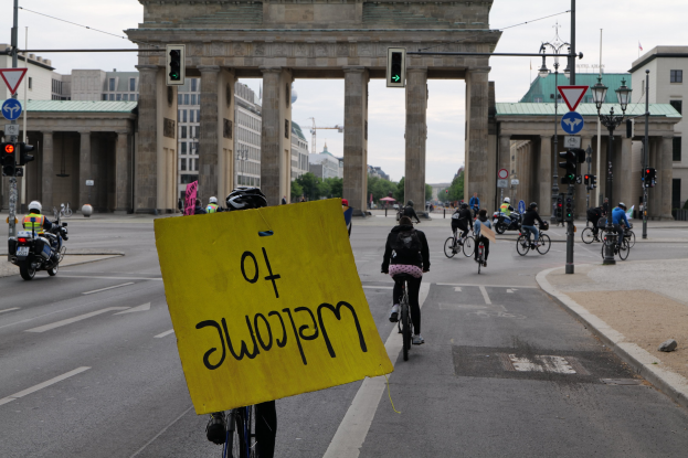 Eine Gruppe von Radfahrern mit Helmen fährt an der Brandenburgertor in Berlin vorbei, einer hält ein gelbes Schild, mit Laternenmasten, Ampeln, Gebäuden, Bäumen und einem klaren blauen Himmel im Hintergrund.