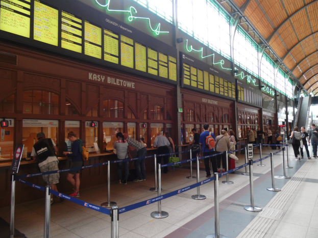Eine Gruppe von Menschen steht in einer Schlange an einem Bahnhof, einige tragen Taschen, mit Pfählen, die mit Bändern geschmückt sind, Schildern, Deckenlampen und Treppen mit Geländern, und einem Gebäude im Hintergrund.