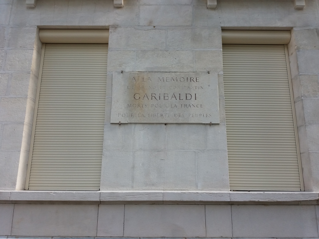 Ein zweistöckiges Gebäude mit zwei Fenstern, jeweils mit Läden, und einer Gedenktafel an der Seite.