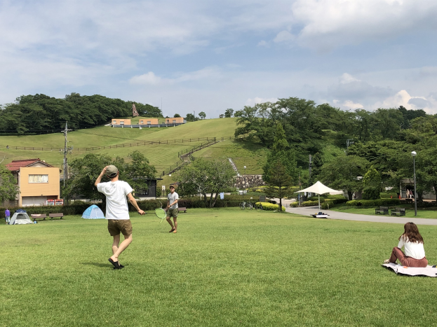 Gruppe von Menschen, die Badminton in einem Park spielen, mit einem Mann, der einen Schläger hält und auf Gras in der Nähe von Zelten und Gebäuden sitzt, mit Hügeln und bewölktem Himmel im Hintergrund.