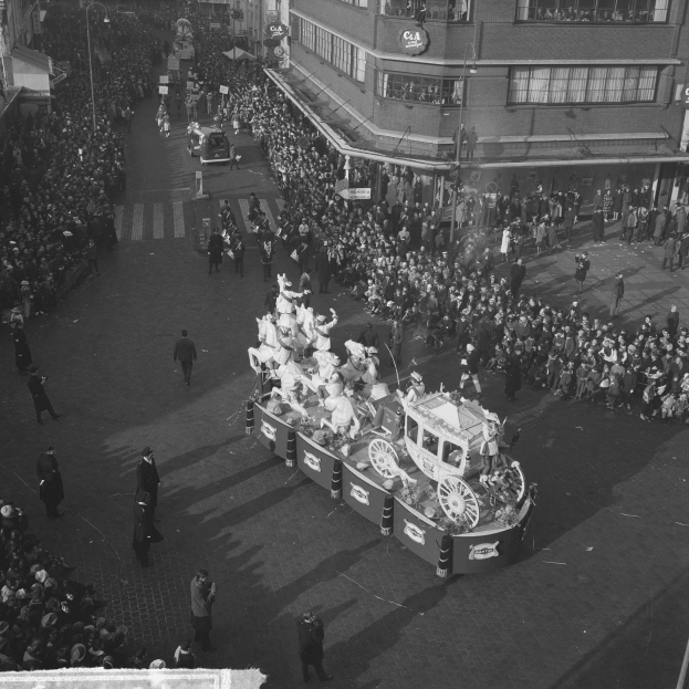Eine große Menschenmenge umringt einen Schmuckwagen bei einem Umzug, dekoriert mit bunten Luftballons und Bändern, mit Gebäuden und Laternen im Hintergrund, in Schwarzweiß.