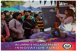 Gruppe von Menschen um einen Tisch versammelt mit einem "Alumni Engagement Innovation Fund"-Schild, umgeben von Gegenständen, mit Zelten und Bäumen im Hintergrund unter einem klaren blauen Himmel.